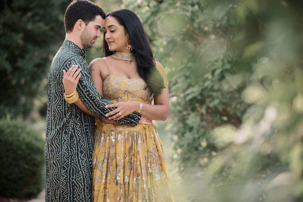 Couple in traditional South Asian engagement attire embracing outdoors with ivy-covered wall backdrop