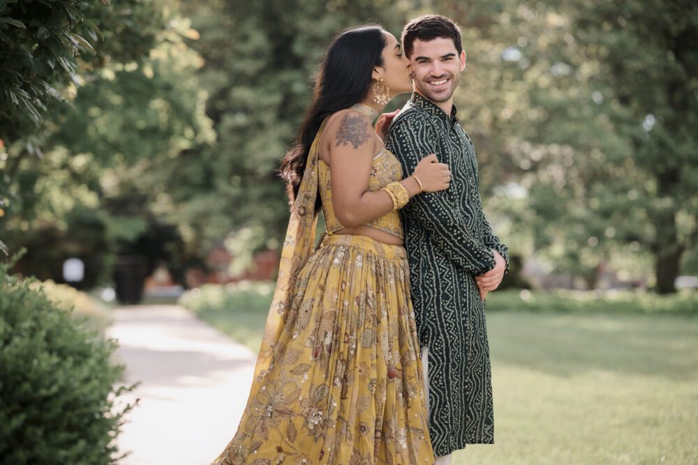Couple in traditional South Asian engagement attire posing together in a tree-lined garden setting