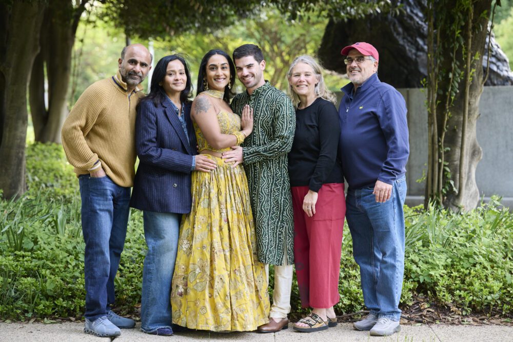 Six adults smiling at camera in tree-lined garden path, wearing casual and traditional Indian attire