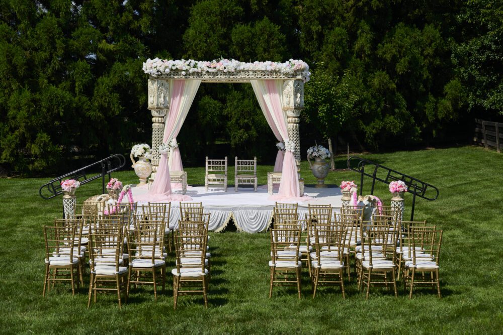 Outdoor wedding ceremony setup on manicured lawn with ornate white altar, pink and white floral arrangements, and gold Chiavari chairs.