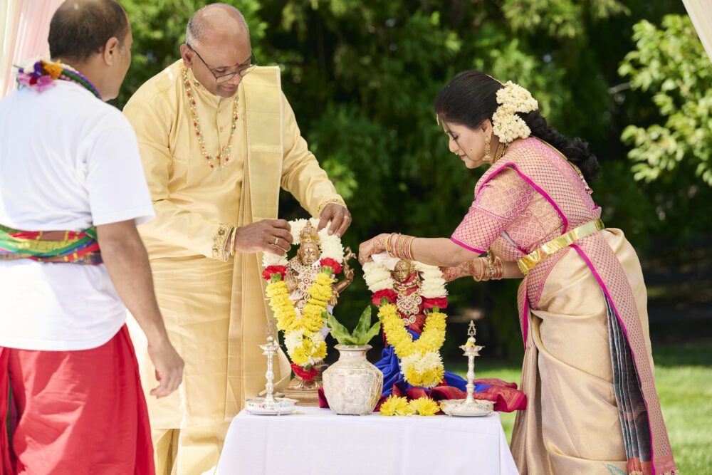 Man in yellow dhoti and woman in pink silk saree performing puja ritual at decorated altar with flower garlands