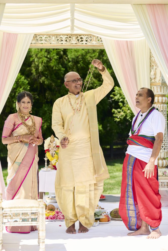 Man in yellow sherwani raising his hand during a Shashtipoorthi ceremony under an ornate white pavilion with pink and cream draping